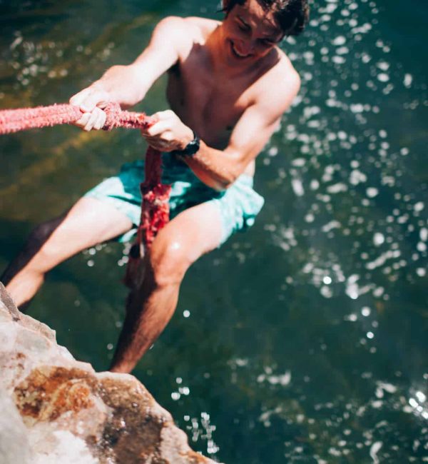Man climbing rock by water with a rope, outdoor adventure.